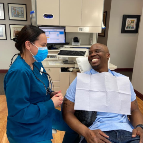 A female dental technician conversing with a smiling male African-American patient in a dental treatment chair.